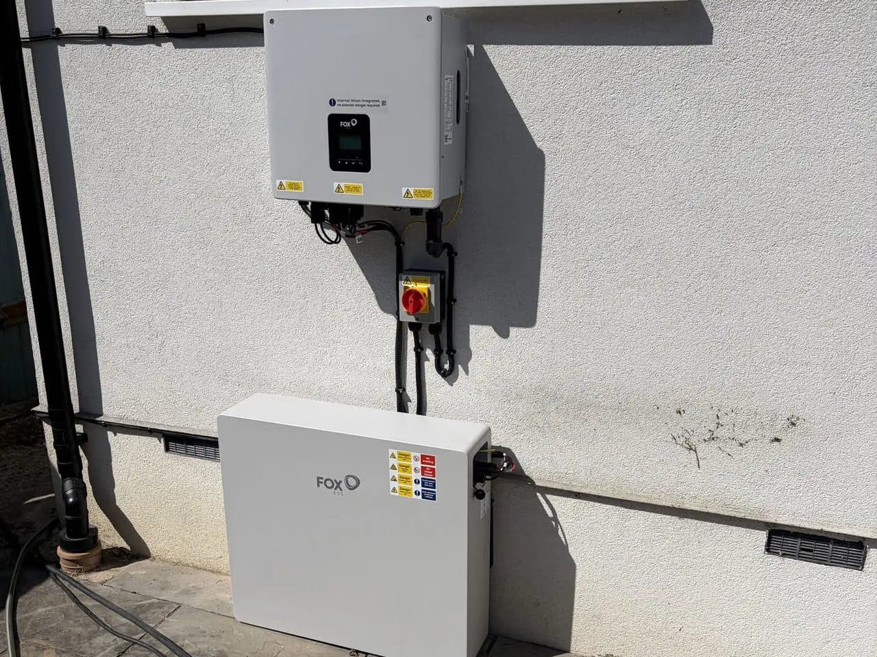 A FoxESS hybrid inverter mounted on an outside wall above a FoxESS battery standing on the patio.
