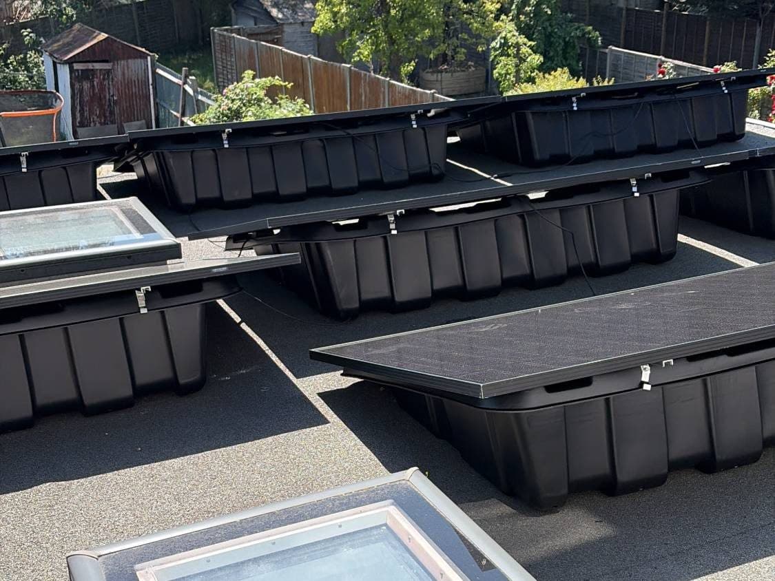 Black solar panels mounted in weighted frames on a flat roof, with gardens behind.